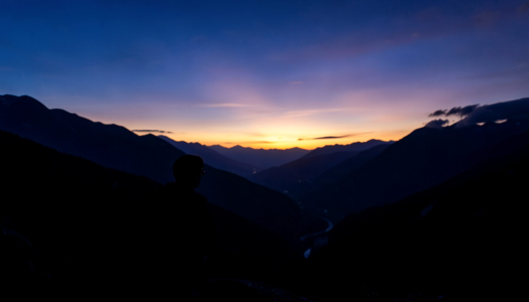 Silueta de una persona contemplando un valle al amanecer, tonos azules, morados y dorados, atmósfera contemplativa.