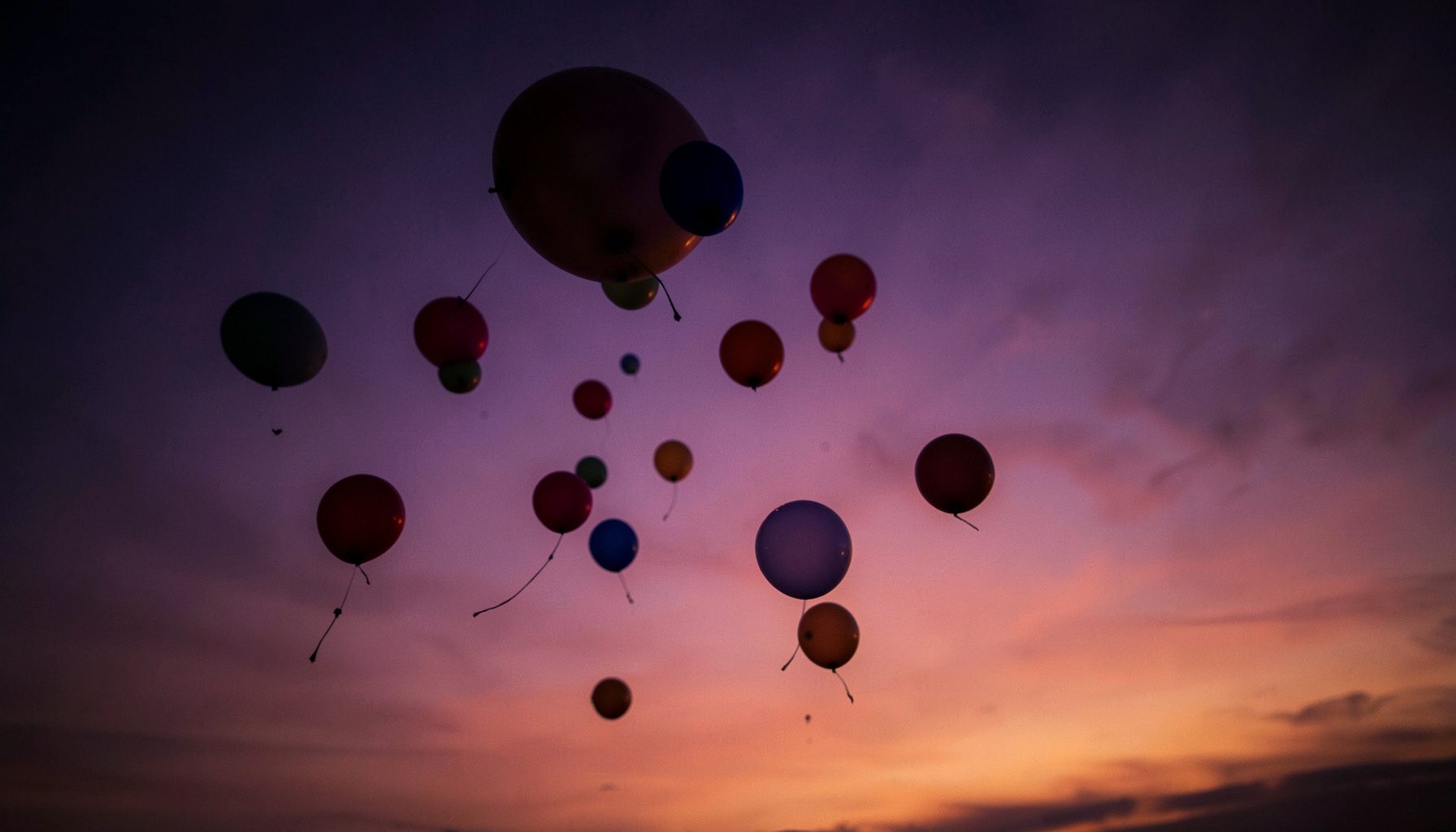 Globos de colores cálidos elevándose contra un cielo crepuscular en tonos violeta y naranja, sensación de libertad y alegría.